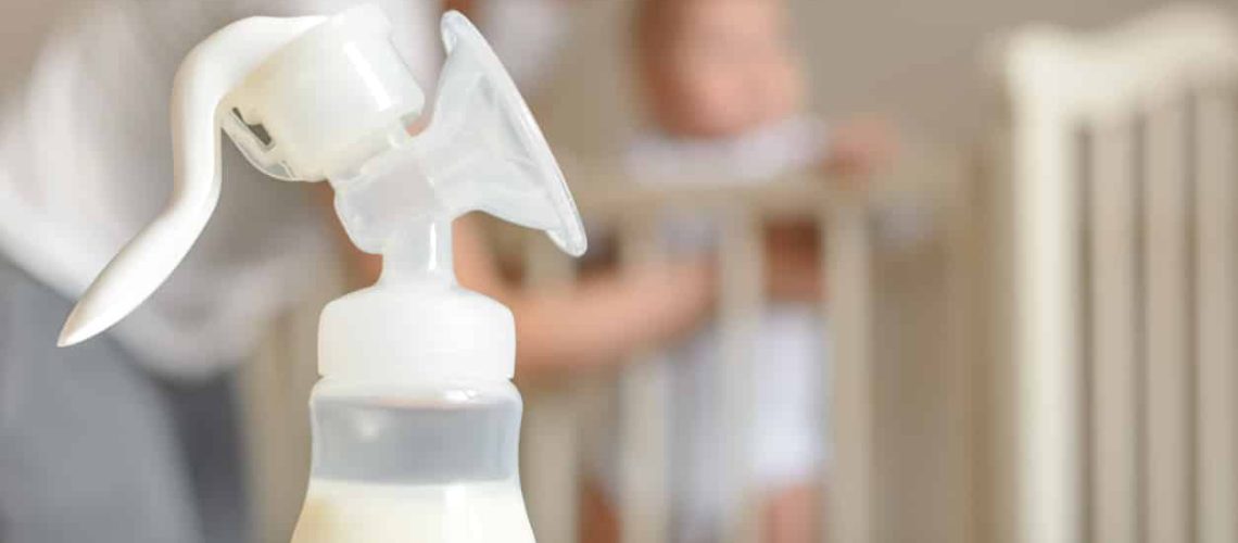 Manual breast pump and bottle with breast milk on the background of mother and baby near the baby's bed.